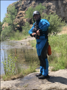 Kody Simons, standing on a riverbank in full rescue gear, leading a class on swift water rescue.